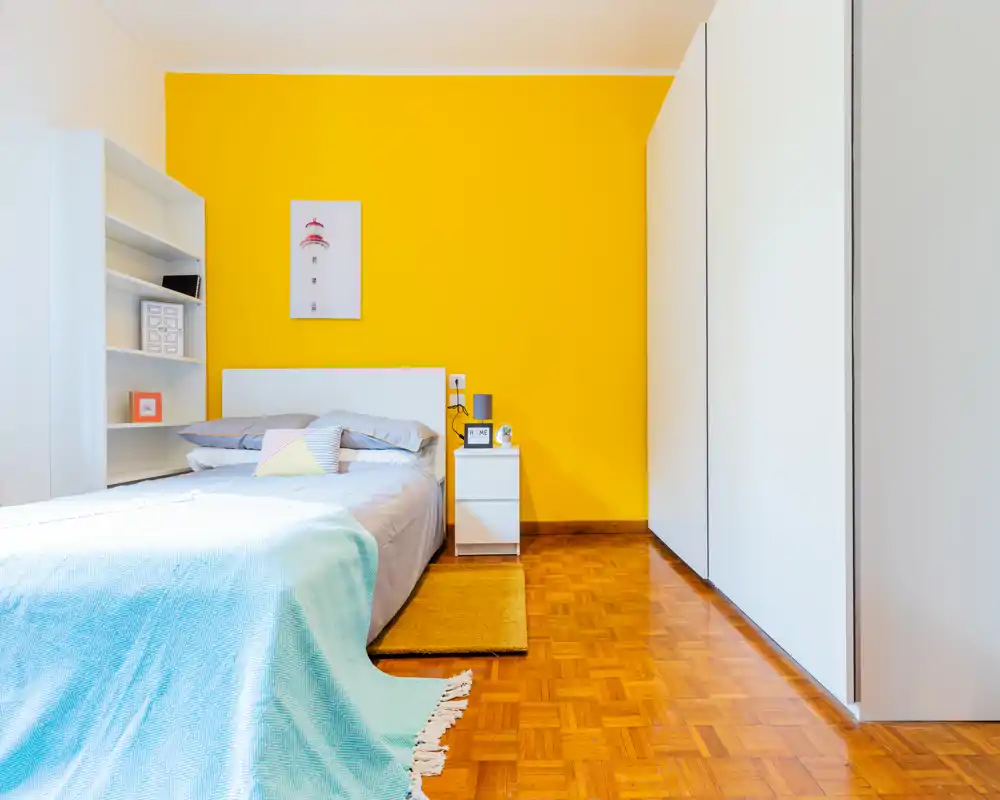 Camera da letto luminosa e moderna con letto bianco su una parete gialla vivace, comodino e armadio a muro. Pavimento in legno, biancheria ordinata e cuscini decorativi creano un ambiente fresco e accogliente, perfetto per annunci.