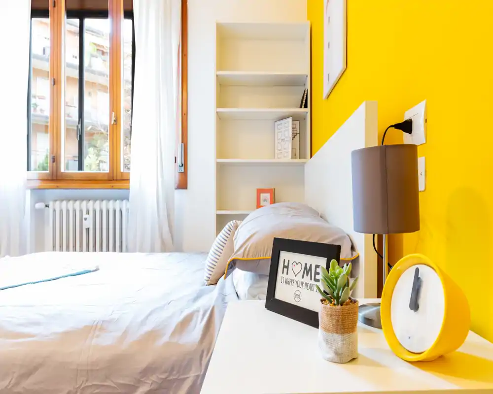 Camera da letto luminosa e moderna con letto matrimoniale ben rifatto, parete gialla, comodino con lampada e orologio e una finestra ampia che lascia entrare luce naturale.