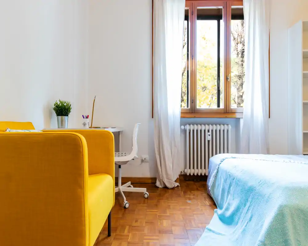 Camera da letto luminosa e ordinata con letto con biancheria chiara accanto a una grande finestra; una poltrona gialla e una scrivania creano un angolo studio accogliente.