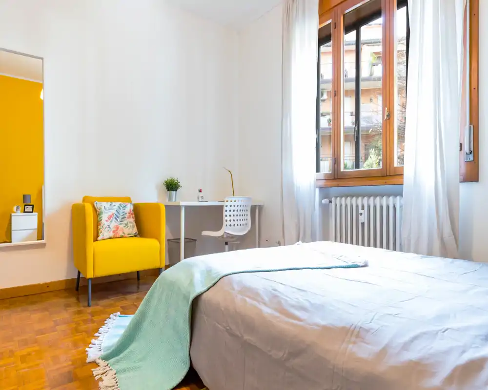 Camera da letto luminosa e ariosa con un comodo letto matrimoniale, una poltrona gialla accogliente e una scrivania semplice vicino alla finestra — ideale per riposo e lavoro leggero.
