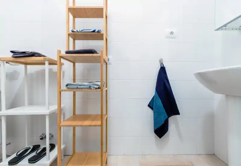 Bright, clean bathroom corner with tiled walls, a pedestal sink and wooden shelving holding towels and slippers.