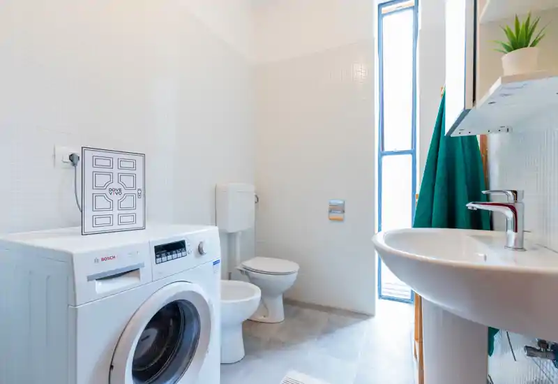 Clean, bright bathroom with a washing machine, toilet, bidet and pedestal sink; white tiles and natural light from a tall window create an airy feel.