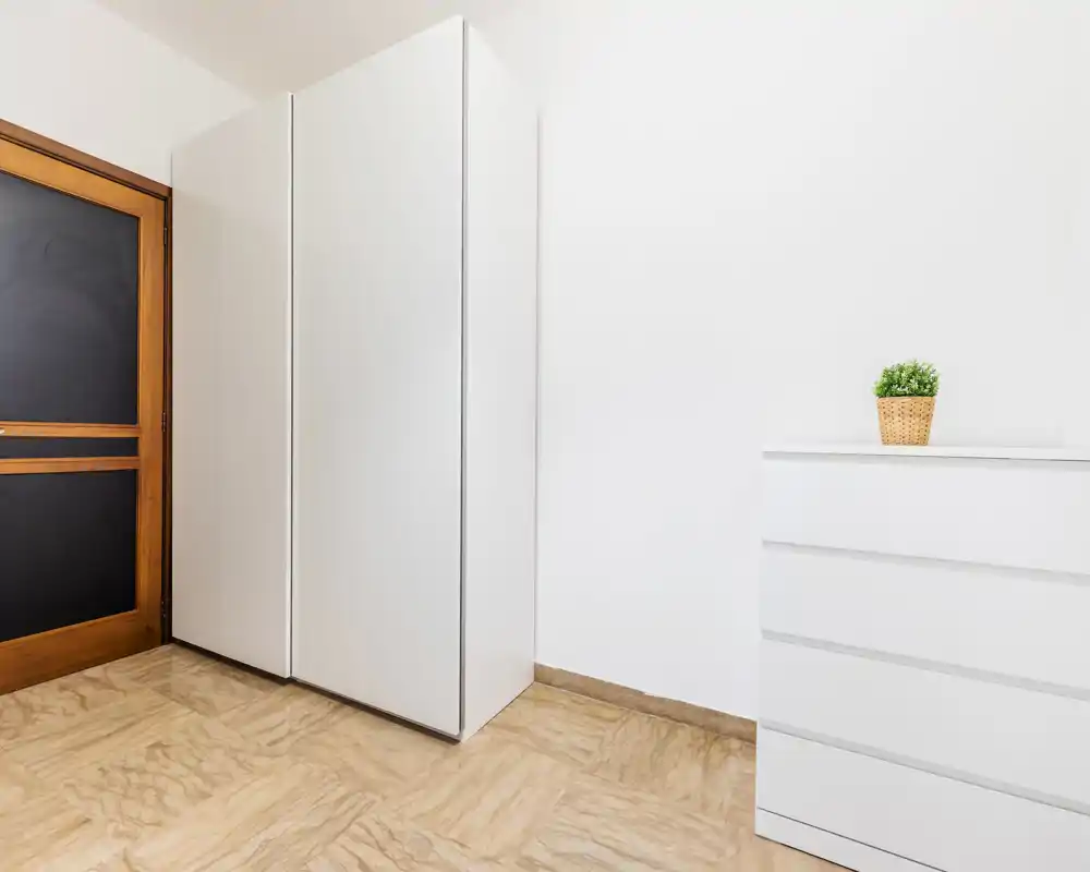 Coin de pièce minimaliste et lumineux avec armoires blanches et petite commode ; espace simple et bien éclairé, idéal pour rangement ou entrée mise en scène.
