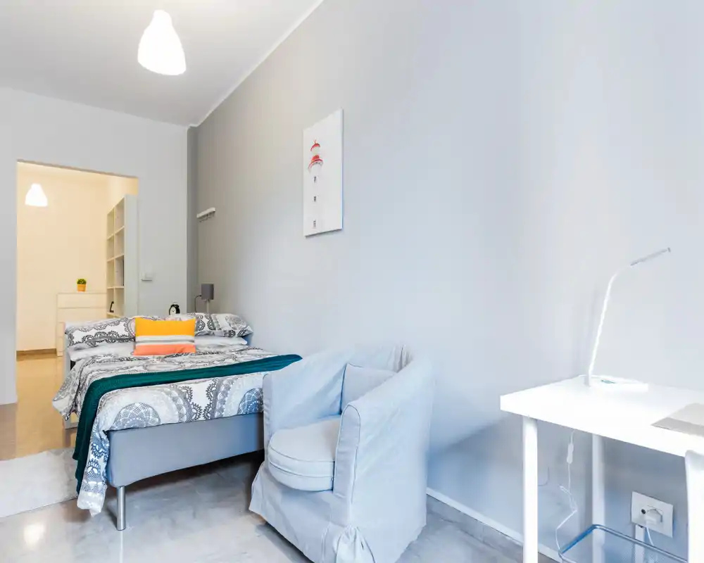 Chambre lumineuse et moderne avec un lit double bien fait, coussins décoratifs et un fauteuil confortable. Décoration minimaliste et murs neutres qui apportent calme et convivialité.