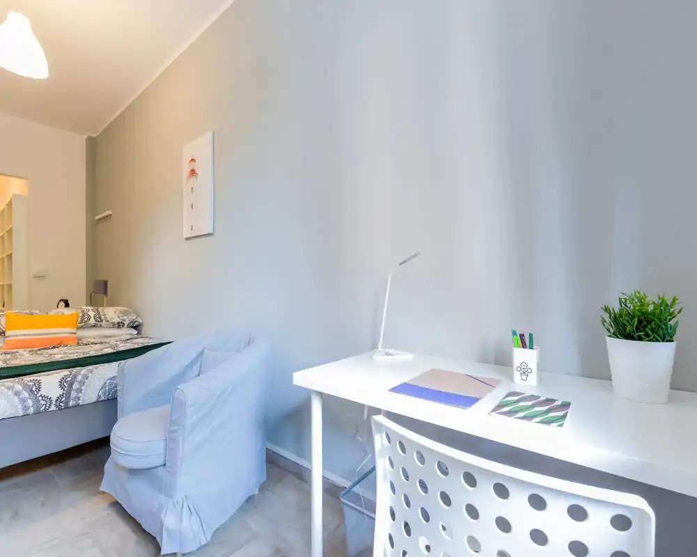 Chambre lumineuse et bien mise en scène avec un lit confortable, un fauteuil moelleux et un bureau blanc. Les tons neutres et les coussins décoratifs apportent une ambiance calme et accueillante, idéale pour les annonces.