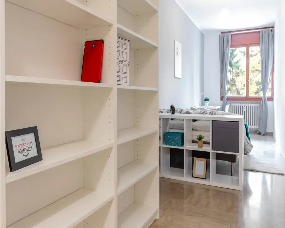Chambre lumineuse et minimaliste avec le lit partiellement visible derrière une étagère ouverte ; une grande fenêtre et une déco sobre donnent une atmosphère calme.