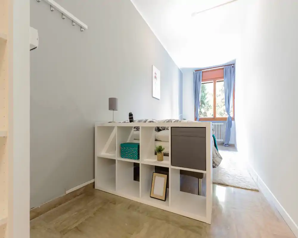 Chambre compacte et lumineuse avec un meuble de rangement au pied du lit, textiles doux et une fenêtre apportant la lumière naturelle.
