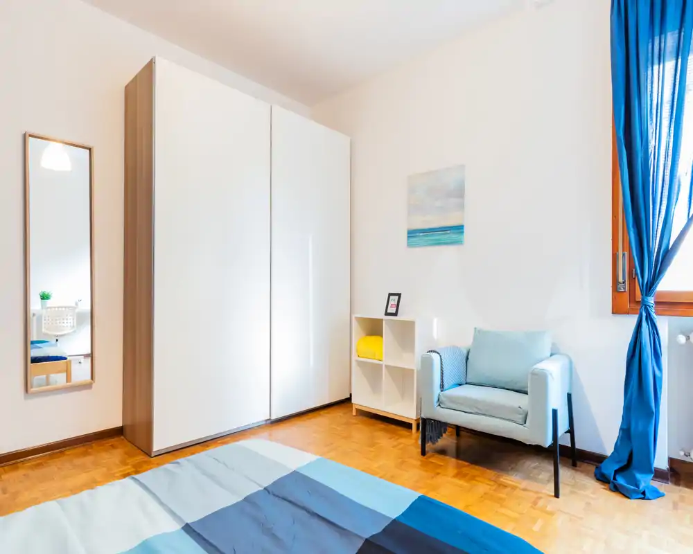 Camera da letto luminosa e moderna con copriletto blu, una poltrona confortevole, ampio armadio e luce naturale dalla finestra — ideale per un soggiorno rilassante.