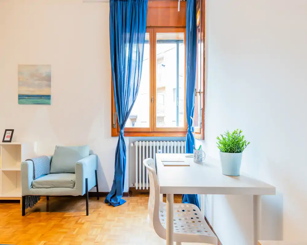 Zona giorno luminosa e ariosa con una poltrona confortevole, una scrivania ordinata vicino alla finestra e gradevoli accenti blu — ideale per rilassarsi o lavorare.