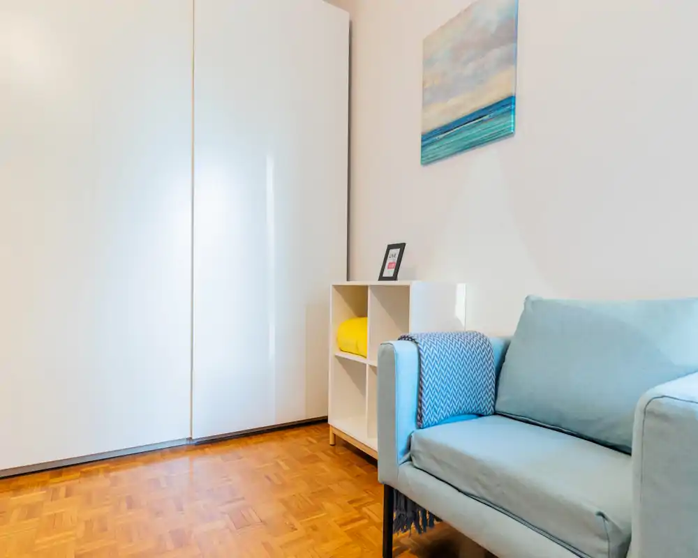 Zona giorno compatta e luminosa con un divano azzurro chiaro, piccolo mobile a cubi e un grande armadio bianco; pavimento in parquet e quadro a tema marino creano un'atmosfera fresca e minimal.