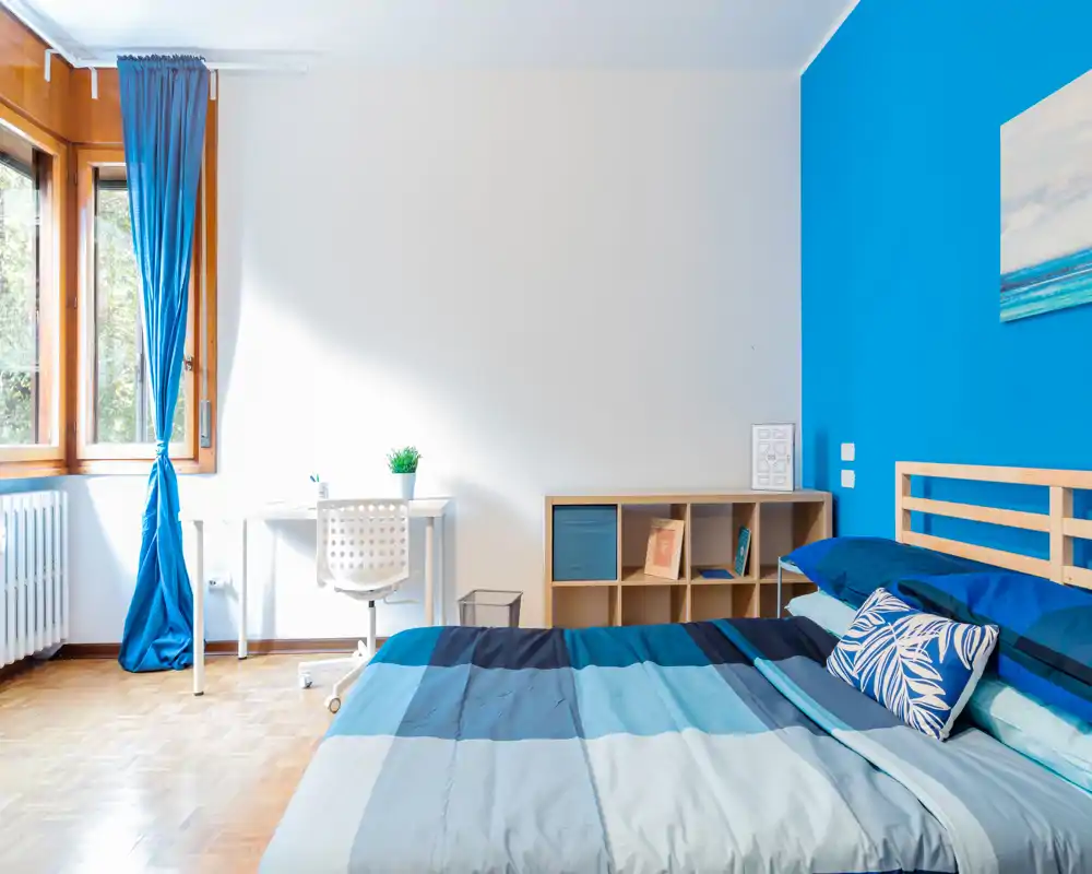 Camera da letto moderna e luminosa con un ampio letto con biancheria a righe blu, testiera in legno e cuscini coordinati; scrivania e scaffale vicino alla finestra soleggiata rendono lo spazio funzionale e di design.