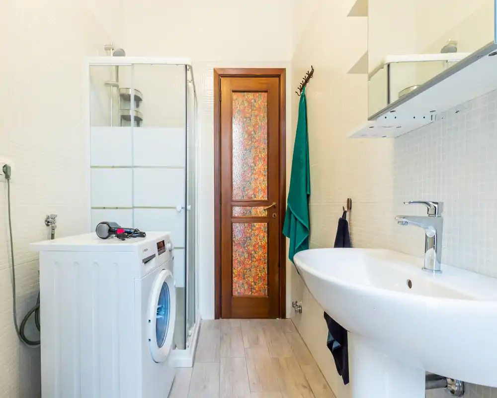 Bagno luminoso e moderno con lavabo a colonna, cabina doccia e lavatrice; piastrelle chiare e pavimento effetto legno creano uno spazio pulito e funzionale.