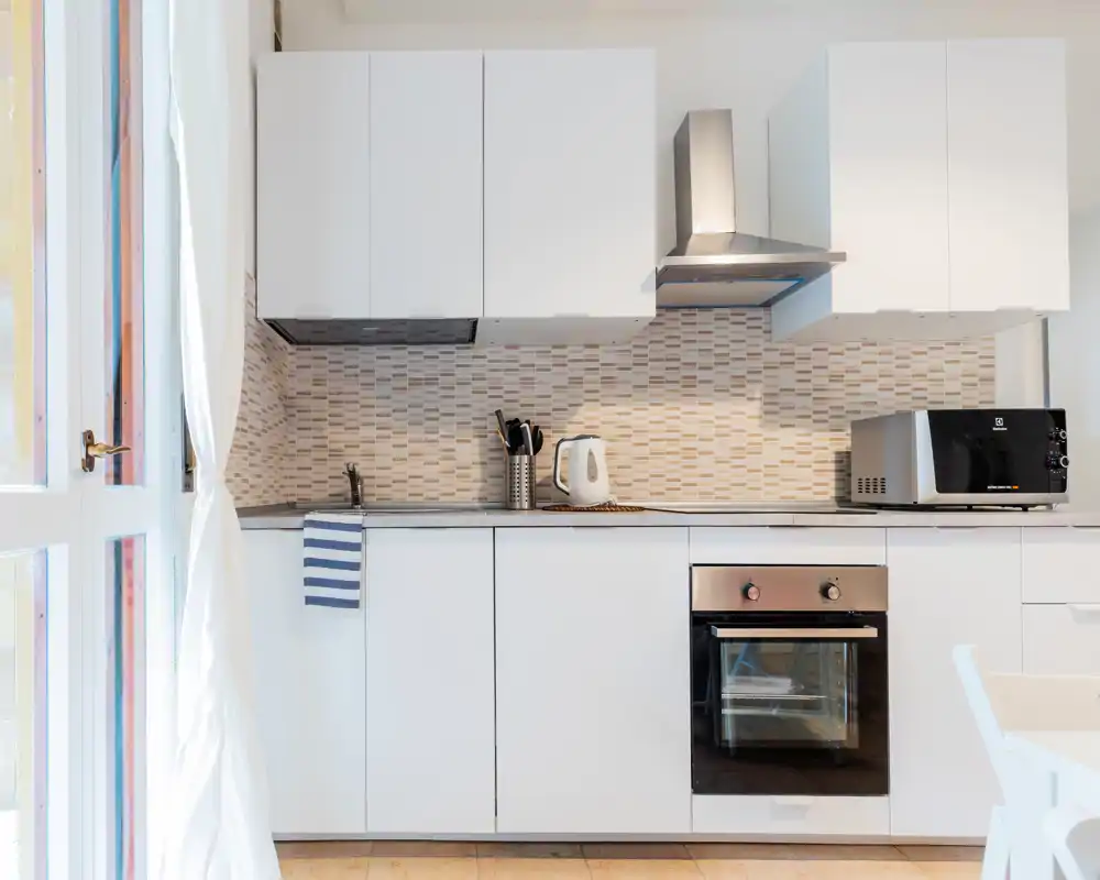 Cucina moderna compatta con mobili bianchi, schienale piastrellato e forno integrato. La luce naturale dalla porta illumina l'ambiente; presente microonde, bollitore e piano di lavoro ordinato.