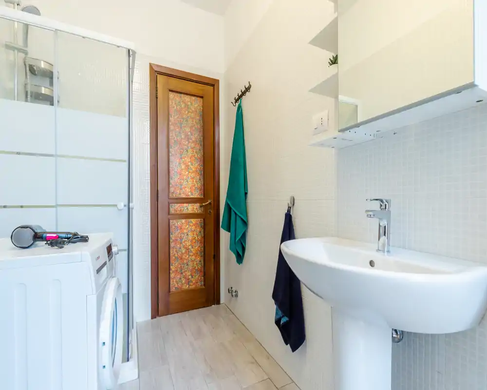 Bagno compatto e luminoso con lavabo a colonna, specchiera a muro, ganci per asciugamani e box doccia parzialmente visibile; piastrelle a mosaico chiare e porta in legno per un tocco caldo.