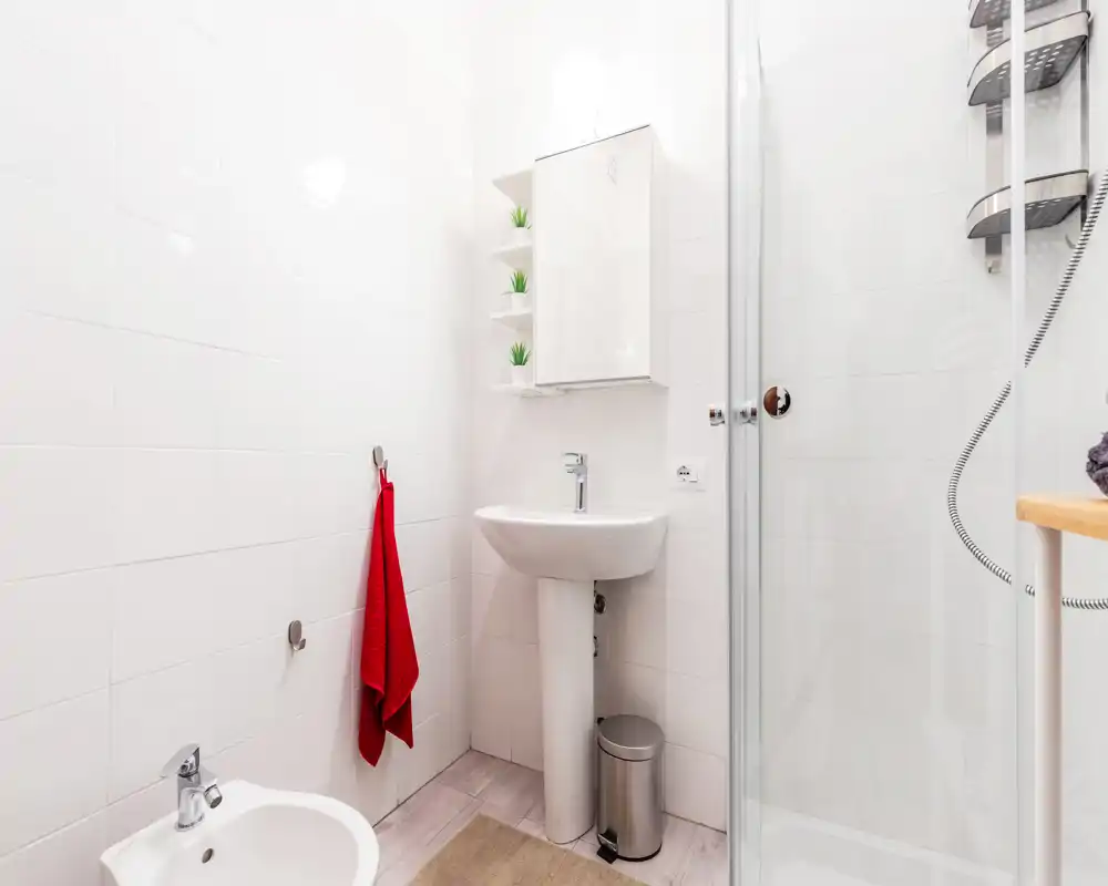 Piccolo bagno moderno con piastrelle bianche, lavabo a colonna, bidet, box doccia in vetro e un tocco di colore con l’asciugamano rosso — pulito e luminoso.