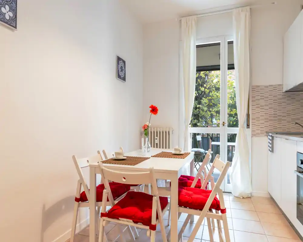 Zona pranzo luminosa e ordinata con tavolo bianco e sedie con cuscini rossi accanto a una piccola cucina aperta e una porta finestra verso il balcone.