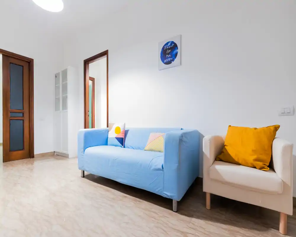 Soggiorno moderno e luminoso con divano coperto di blu, poltrona beige con cuscino senape e pareti bianche pulite—ideale per un soggiorno confortevole.