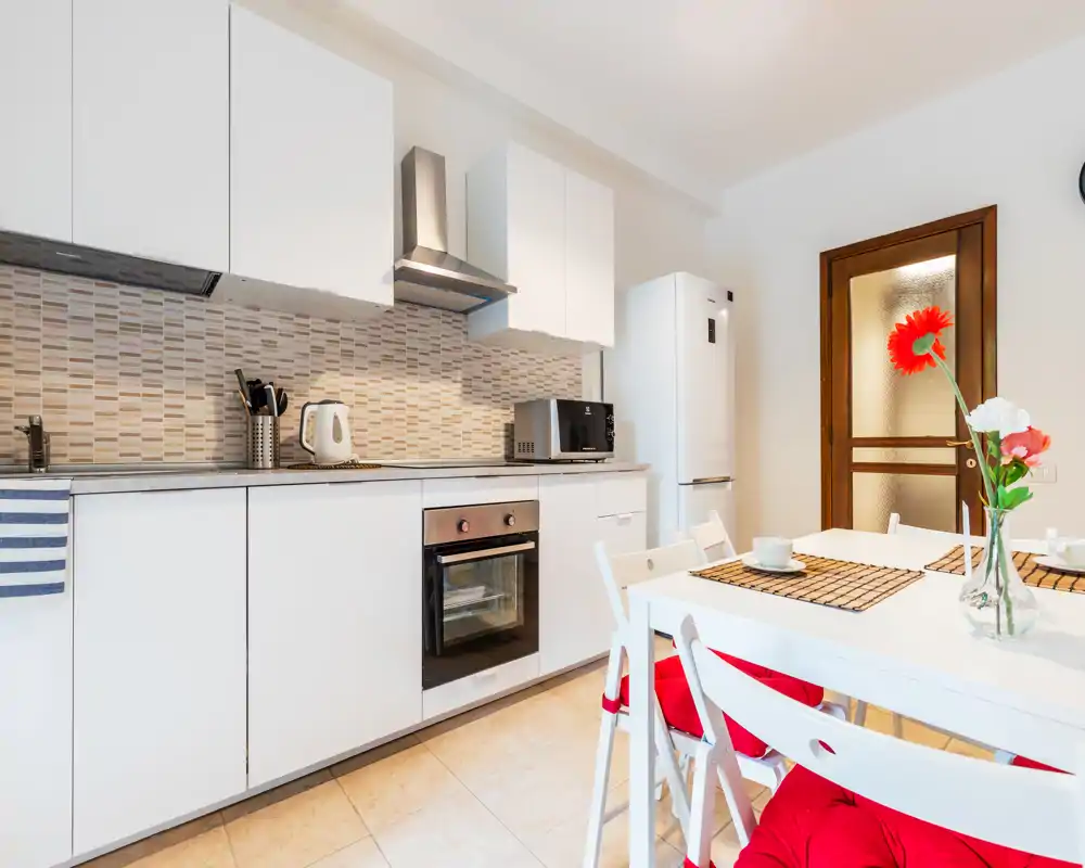 Cucina moderna e luminosa con pensili bianchi, forno integrato, frigorifero e un piccolo tavolo da pranzo con cuscini rossi e fiori freschi — spazio pulito e funzionale ideale per soggiorni brevi.