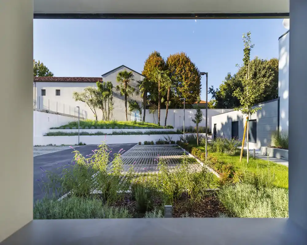 Cortile soleggiato con parcheggi pavimentati, alberi ornamentali e siepi basse, visto attraverso un'apertura architettonica incorniciata. Perfetto per valorizzare le aree comuni esterne e il verde.