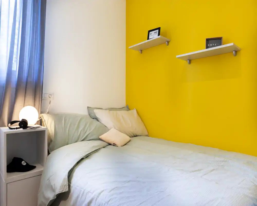 Camera da letto luminosa e ordinata con biancheria dai toni pastello e una parete d'accento gialla vivace; comodino con lampada e mensole semplici per un'atmosfera moderna e accogliente.