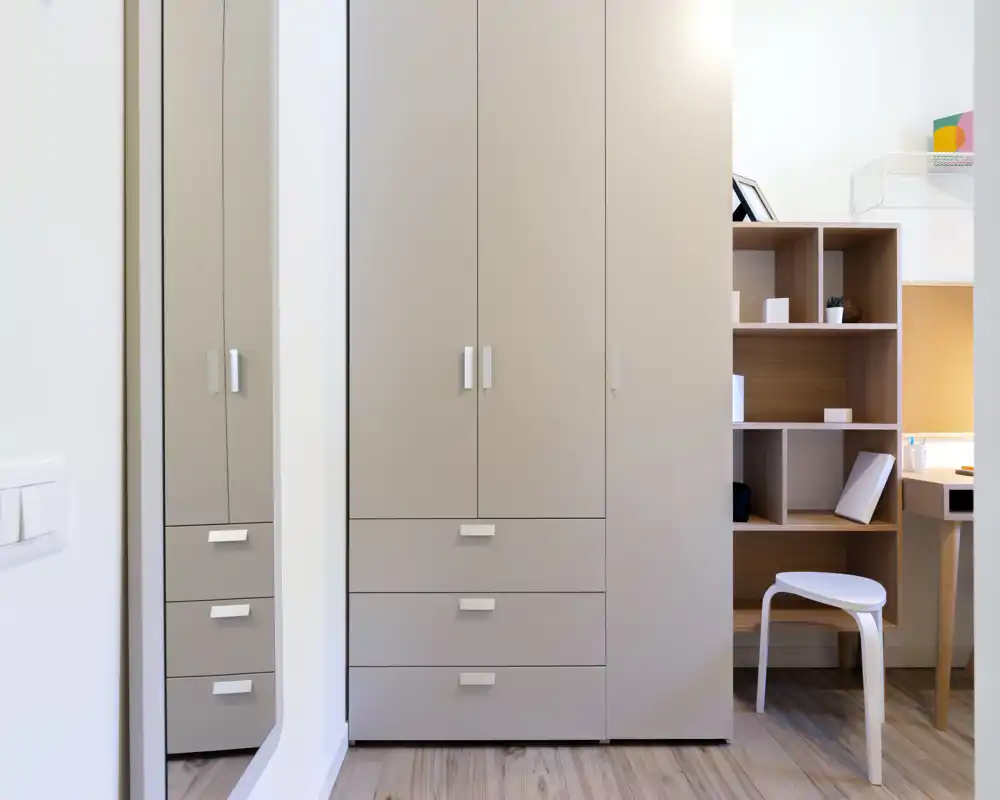 Angolo di camera con armadio alto, specchio e una piccola area scrivania. Finiture moderne e pulite, pavimento in legno chiaro e illuminazione uniforme e luminosa.