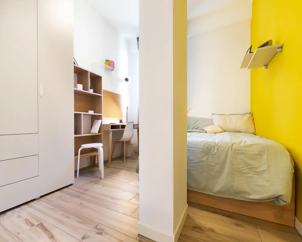 Camera da letto compatta e luminosa con letto singolo contro una parete gialla vivace, biancheria morbida e una piccola mensola. Armadio integrato e scrivania creano uno spazio funzionale e ben illuminato, ideale per uno studente o una persona sola.