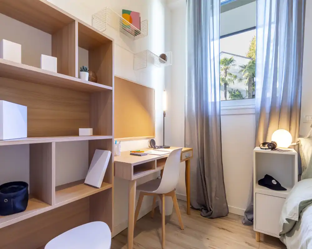 Angolo camera da letto moderno e luminoso con scrivania accanto al letto, tende morbide e scaffalatura elegante — ideale per una stanza in affitto compatta e funzionale.