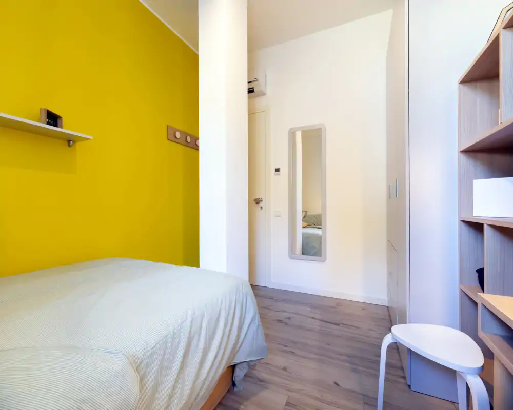 Camera da letto luminosa e ordinata con parete accentuata gialla, letto ben rifatto e arredi semplici. L'ambiente è fresco e ben illuminato, ideale per affitti brevi.