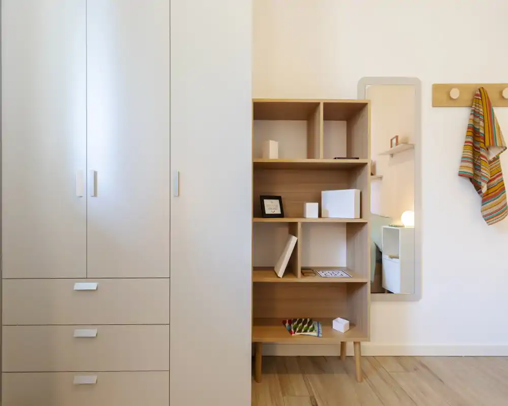 Wardrobe and shelving against a bright wall with a mirror and coat hooks; a tidy, minimal dressing or entry area with light wood flooring and small decorative items.