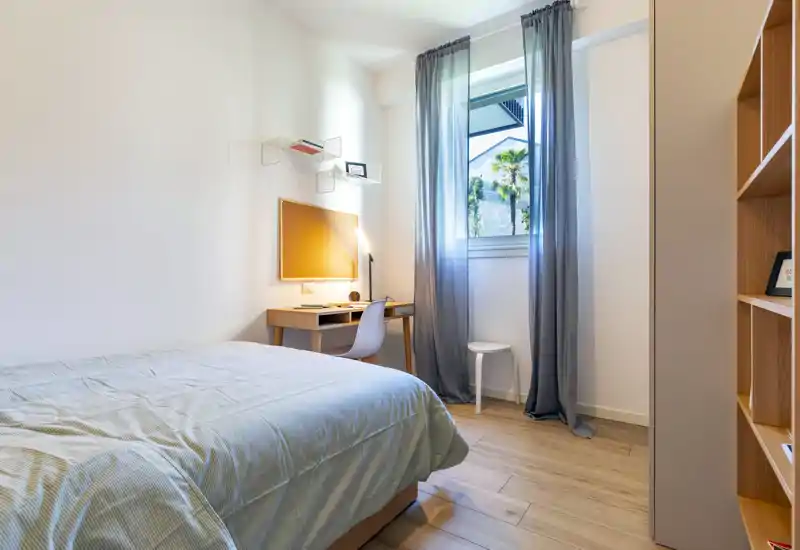 Bright, modern bedroom with a neatly made bed, a small desk and a window letting in natural light — ideal for a comfortable stay or work-from-home.