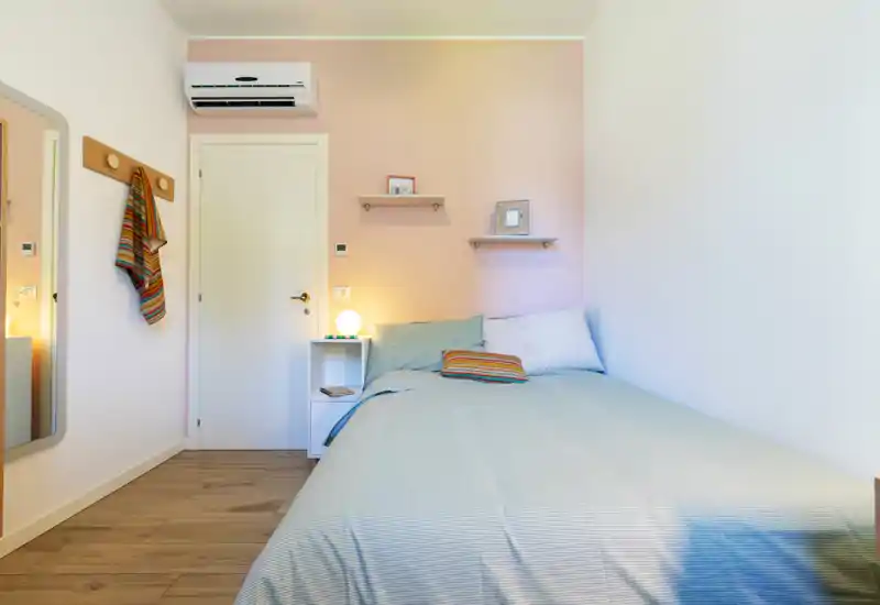 Bright, cozy single bedroom with a neatly made bed, bedside table with lamp, wall shelves and a mirror — ideal for a comfortable short-term stay.