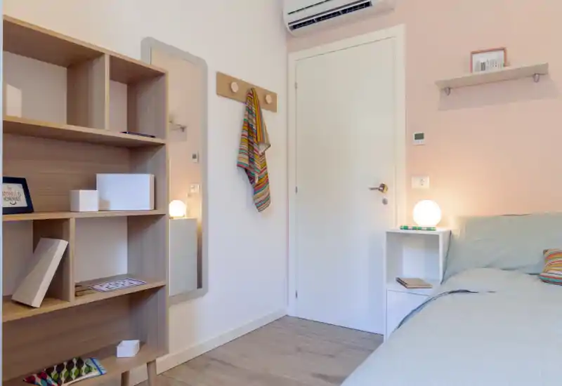 Bright, tidy bedroom corner with a bedside table, lamp and shelving; light pastel walls and soft bedding create a calm atmosphere.