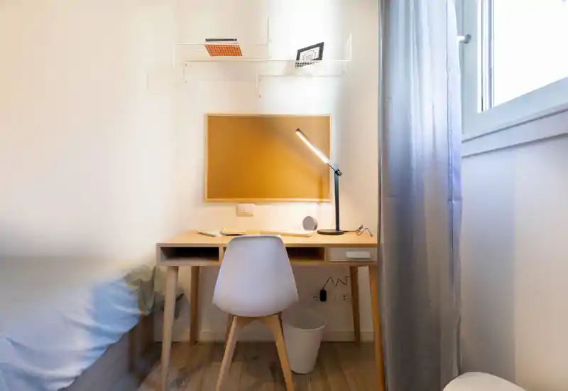 Compact bedroom corner with a minimalist wooden desk and study lamp beside a single bed. Bright natural light from the window and a cork noticeboard make this a practical, tidy student or guest setup.