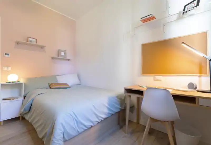 Cozy, well-lit bedroom with a neatly made double bed, bedside table with lamp, and a small desk — ideal for a single occupant or couple seeking a tidy workspace within the sleeping area.