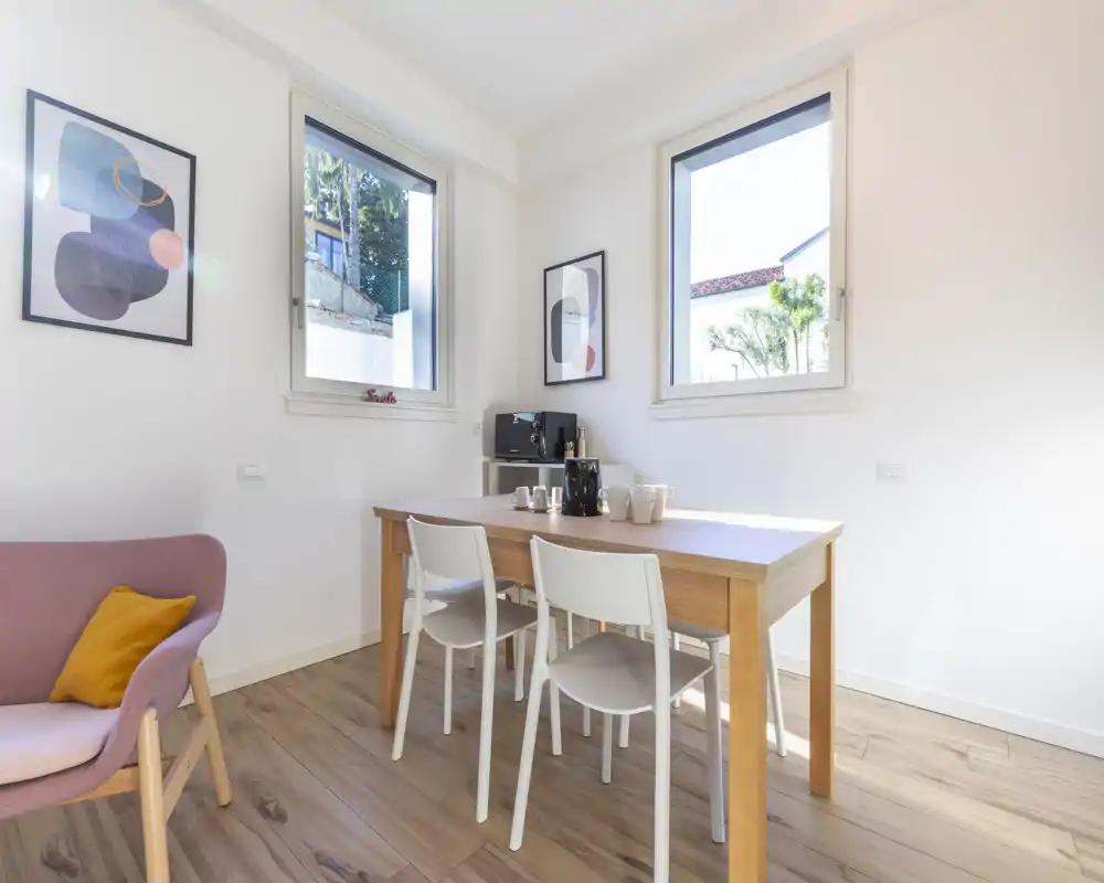 Bright, modern dining area with a wooden table and four white chairs, two large windows providing natural light and simple wall art for a fresh, minimal look.
