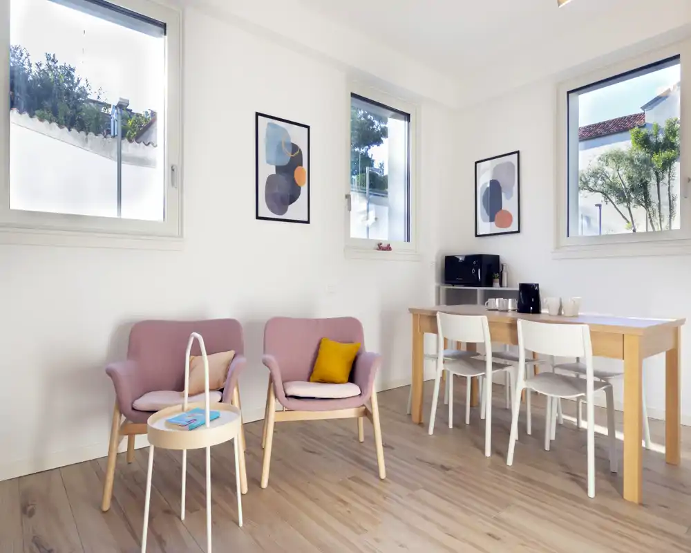 Bright, modern living area with two pink lounge chairs, a small side table and a wooden dining table with white chairs; three windows bring abundant natural light and minimalist framed artwork completes the space.