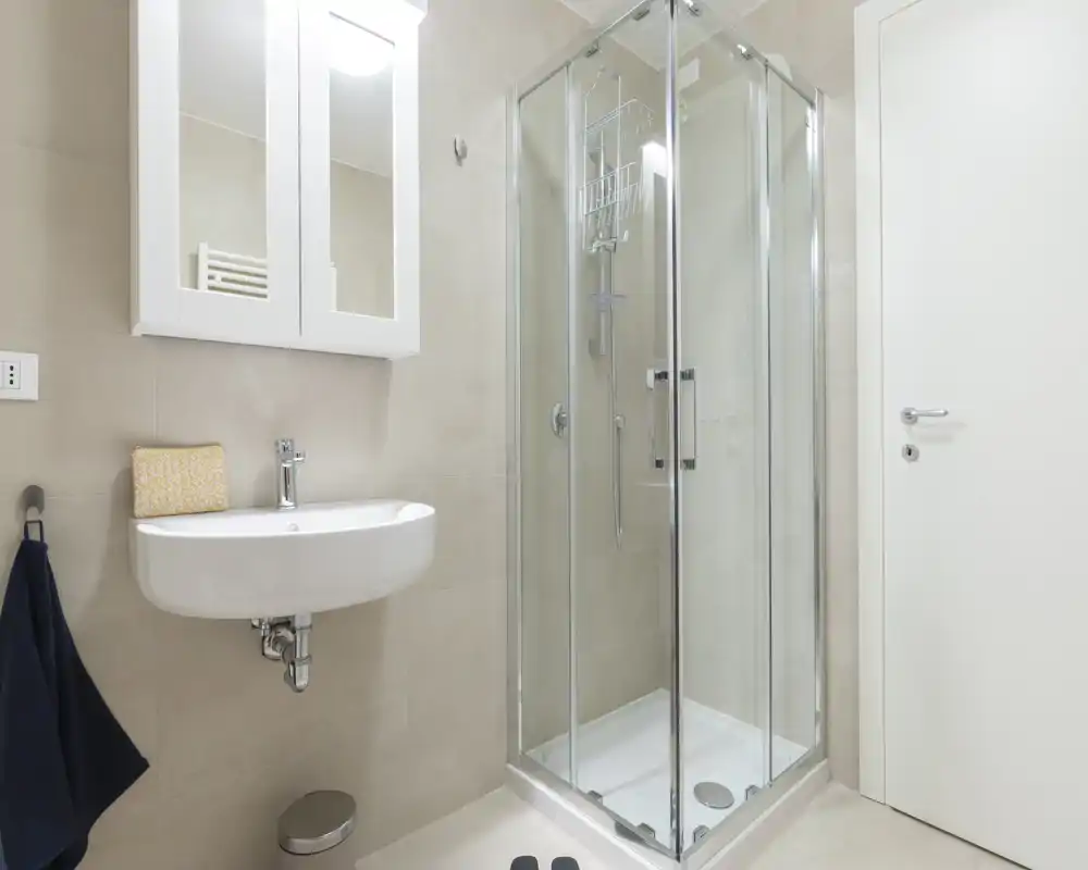 Clean, modern bathroom with a corner glass shower, wall-mounted sink, and mirrored cabinet — bright neutral tiles and simple fixtures.
