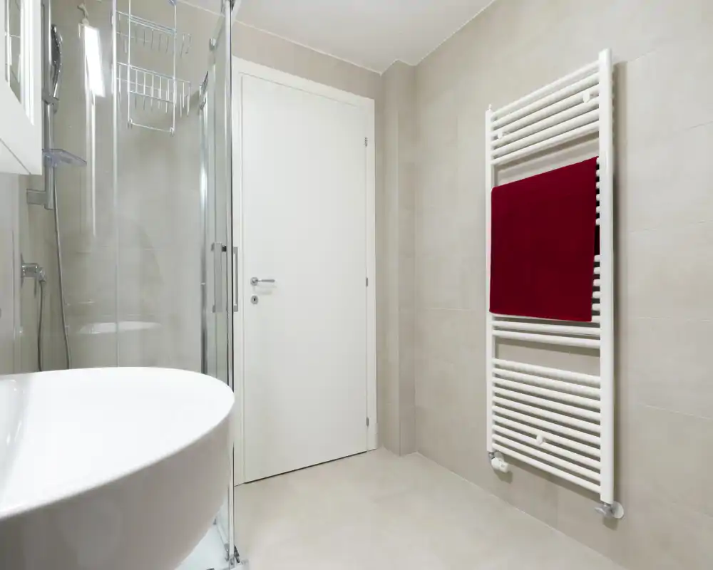 Clean, modern bathroom with a glass shower enclosure, white sink and heated towel rail with a red towel; light neutral tiles create a bright, minimalist space.