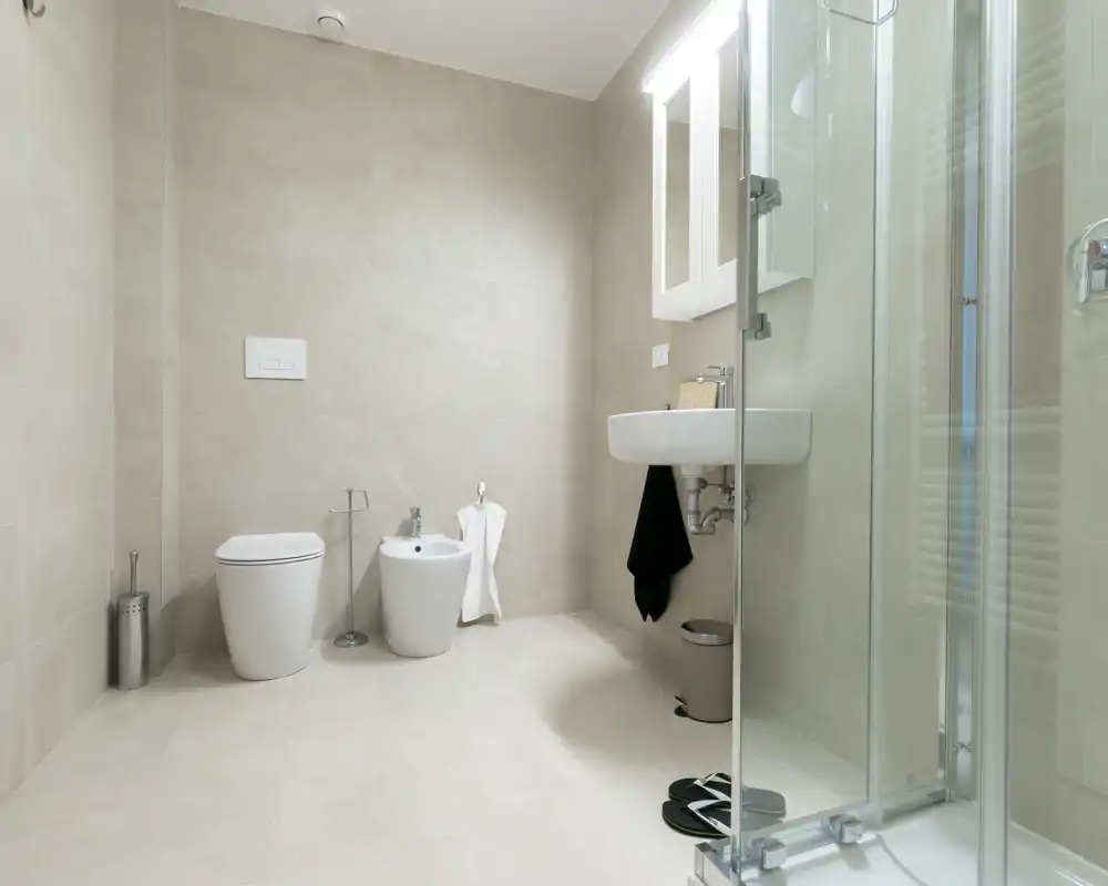 Bright, modern bathroom with wall-mounted sink, toilet, bidet and a glass shower enclosure. Neutral tiles and minimal decor create a clean, spa-like feel.