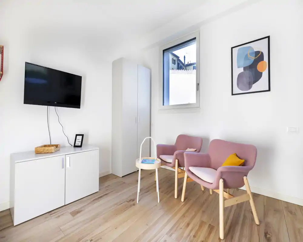 Bright, minimalist seating area with two mauve armchairs, a small side table and wall-mounted TV — ideal for a cozy reading corner in a compact living space.