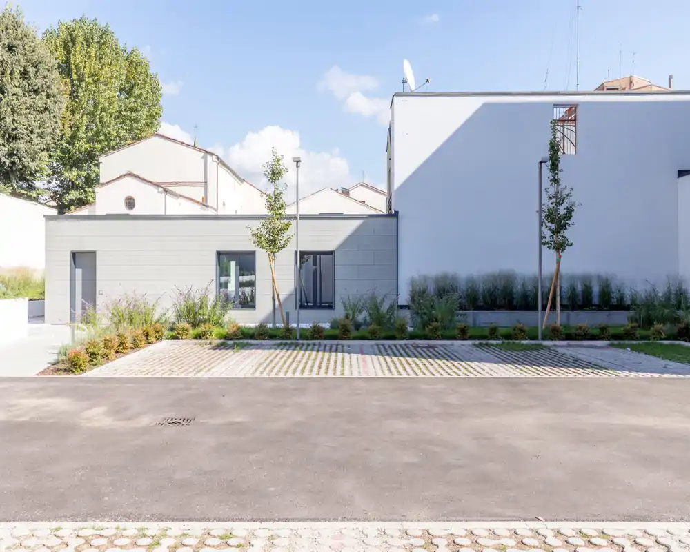 Sunny outdoor parking area with low landscaping: paved parking bays, young trees and a modern low building façade in the background. Bright, tidy exterior suitable for short-term rental photos highlighting available outdoor parking and frontage.