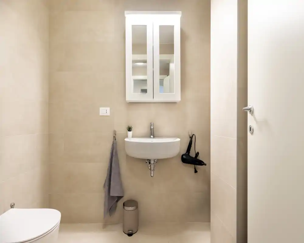 Bright, minimal bathroom with wall-mounted sink, mirrored cabinet, towel hook and a small trash bin — clean, modern neutral tiles.