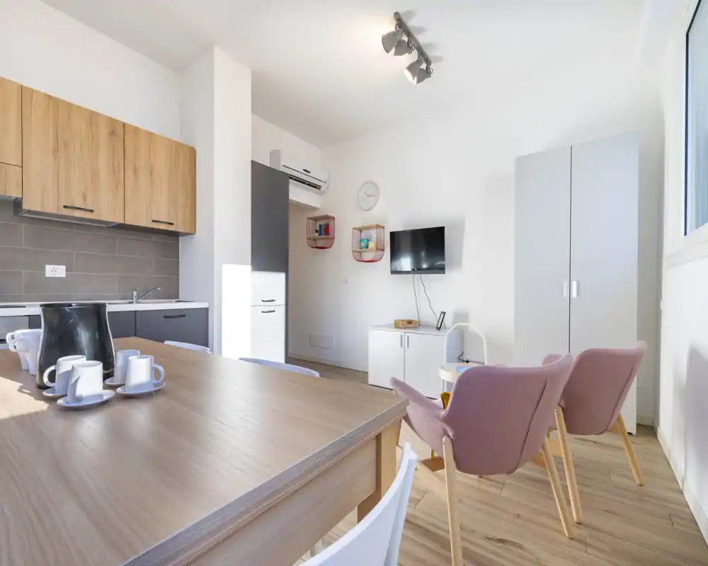 Bright, modern open-plan living area with a small kitchenette, wooden dining table and two comfortable pink armchairs — stylish and well-lit for short stays.