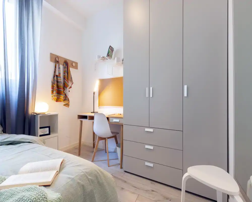 Bright, tidy bedroom with a visible bed, bedside lamp and a compact desk under shelving. Neutral tones and soft textiles create a calm, well-styled space ideal for rest and study.
