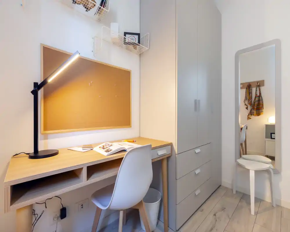 Neat bedroom corner with a study desk, cork noticeboard and wardrobe. Bright, modern styling with a desk lamp and mirror reflecting the room.