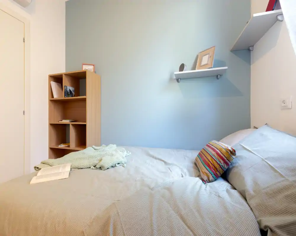 Bright, neatly styled bedroom with a double bed dressed in neutral linens, a colorful cushion and a small shelving unit — ideal for a cozy single or couple stay.