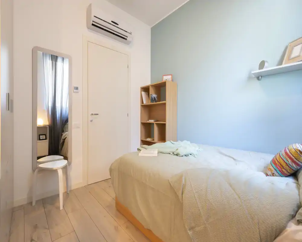 Neat single bedroom with a comfortably made bed, soft linens and warm pastel wall color; a small shelf and mirror create a practical, cozy layout.