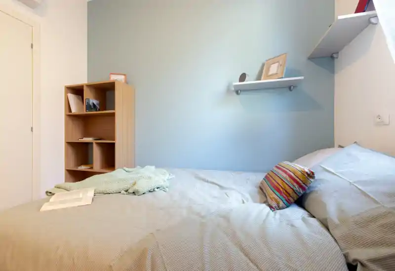 Camera da letto luminosa e ordinata con letto matrimoniale in biancheria neutra, un cuscino colorato e un piccolo scaffale — ideale per un soggiorno accogliente per una o due persone.