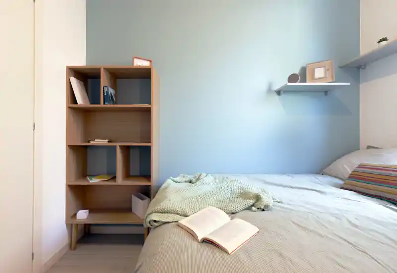 Angolo camera da letto luminoso e ordinato con un letto confortevole, una coperta morbida e un libro aperto sul piumone; una semplice libreria e mensole a parete completano la scena minimal e calma.