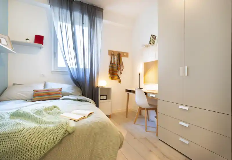 Camera da letto luminosa e accogliente con letto rifatto, coperta lavorata a maglia e una scrivania vicino alla finestra — perfetta come singola o stanza per gli ospiti.